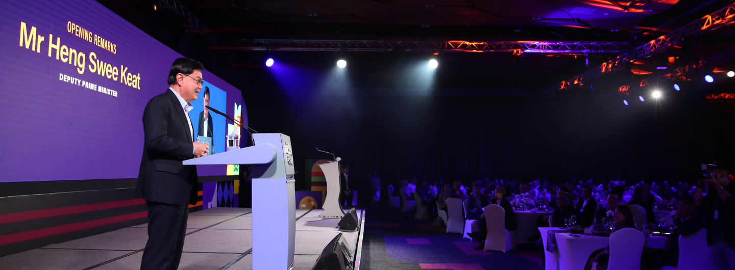 Mr. Heng Swee Keat speaking at a podium, audience seated at round tables. Stage lighting above.